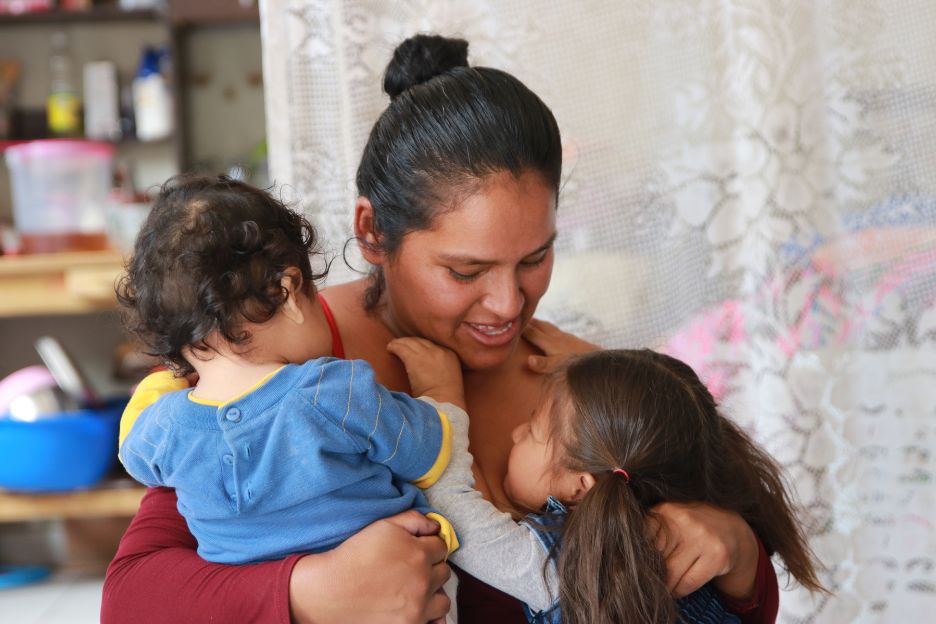 Eine Frau hält zwei kleine Kinder liebevoll in ihren Armen. Die Szene vermittelt eine starke emotionale Bindung und Fürsorge. Im Hintergrund sind Haushaltsgegenstände und ein weißer, gemusterter Vorhang zu sehen, was auf eine häusliche Umgebung hinweist.